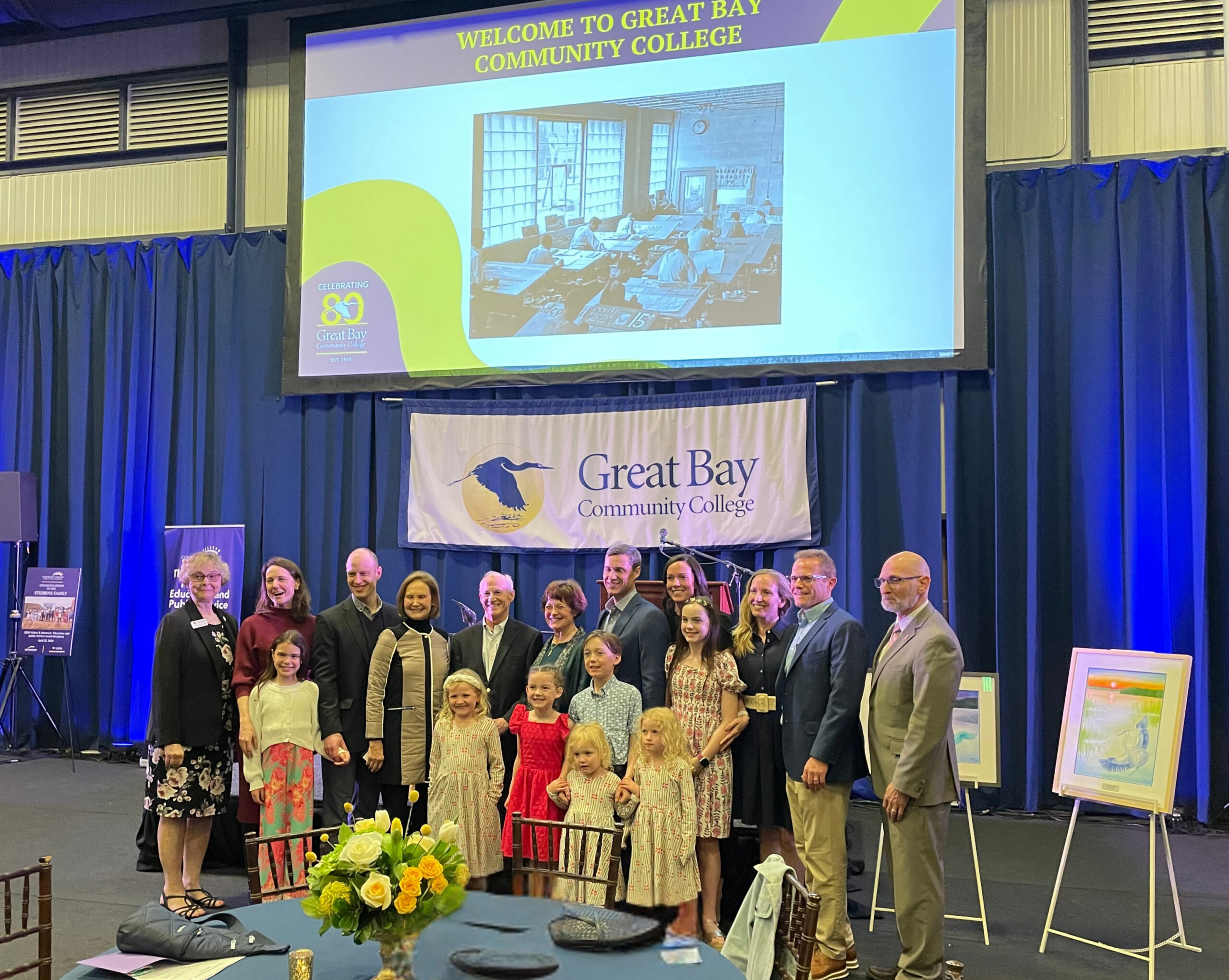 A group poses for a photo at a Great Bay Community College event, standing in front of banners and framed art.