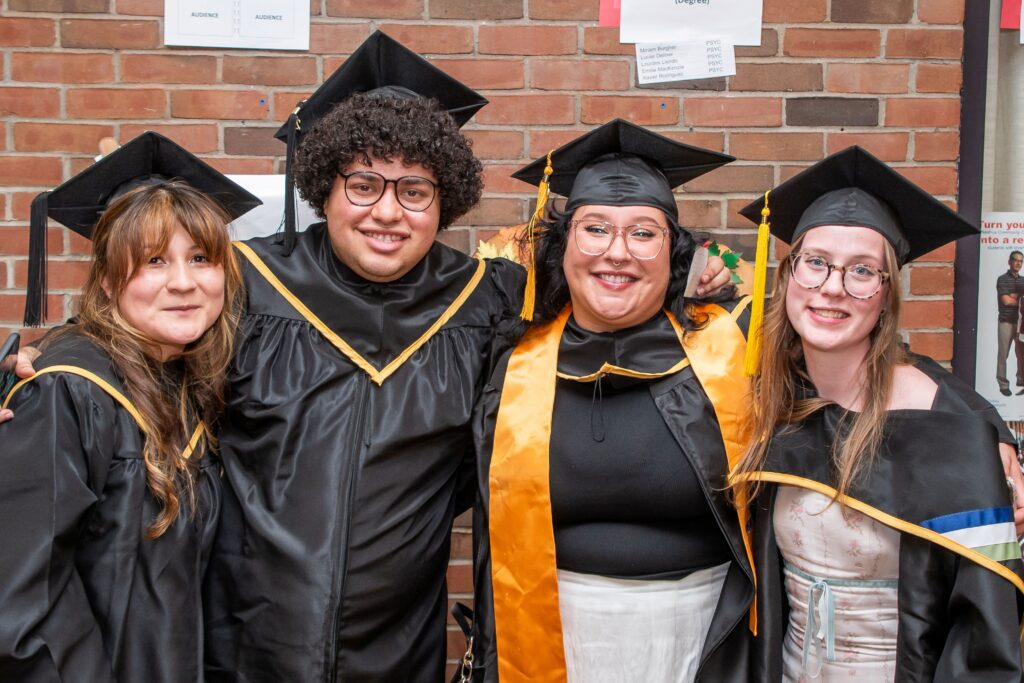 Four graduates in caps and gowns smiling and posing together in front of a brick wall.