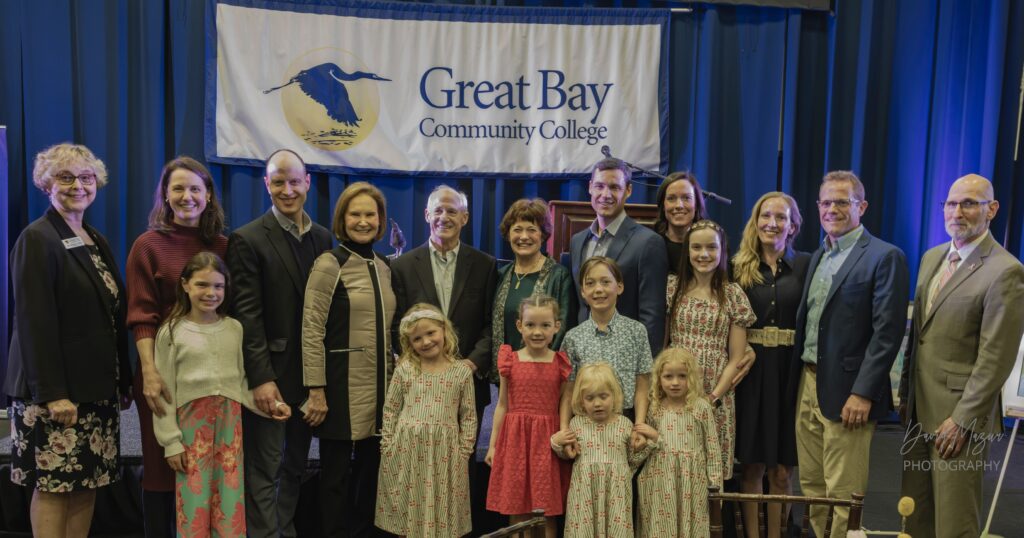 A group of adults and children posing together in front of a Great Bay Community College banner.