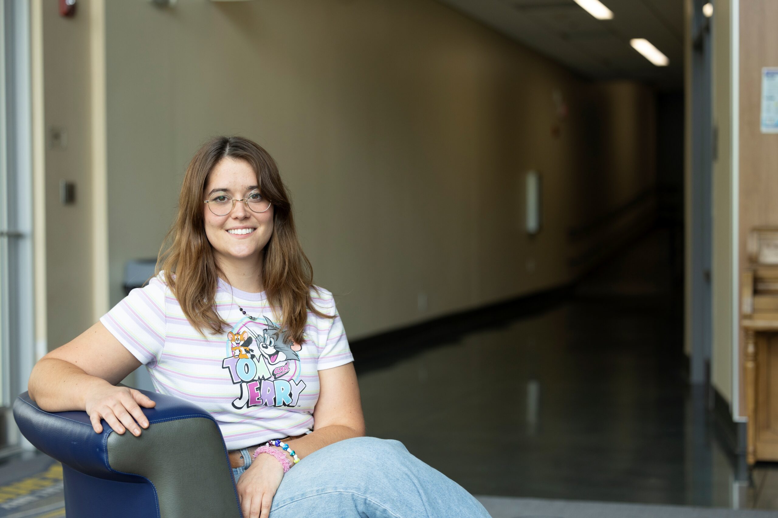 A smiling woman in glasses and a Tom & Jerry shirt sits on a blue chair in a hallway.