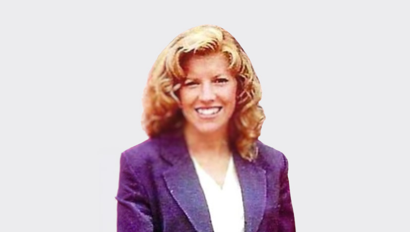Smiling woman with wavy blonde hair wearing a purple blazer and white blouse, against a plain background.