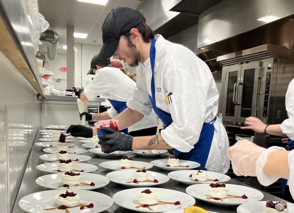Chefs in white uniforms prepare and plate desserts in a professional kitchen, adding sauce to each plate.