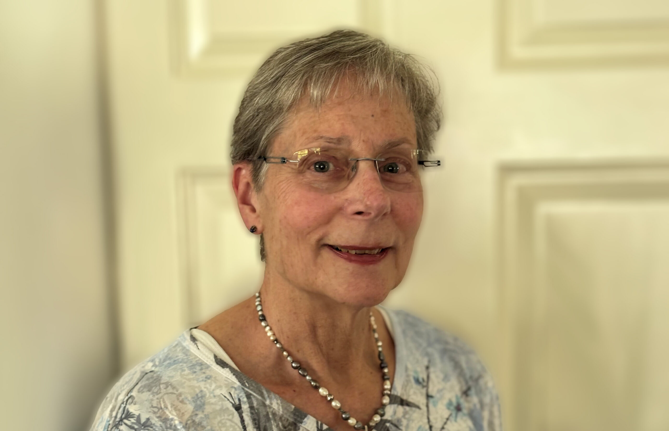 Older woman with short gray hair, glasses, and necklace smiling in front of a cream-colored door.