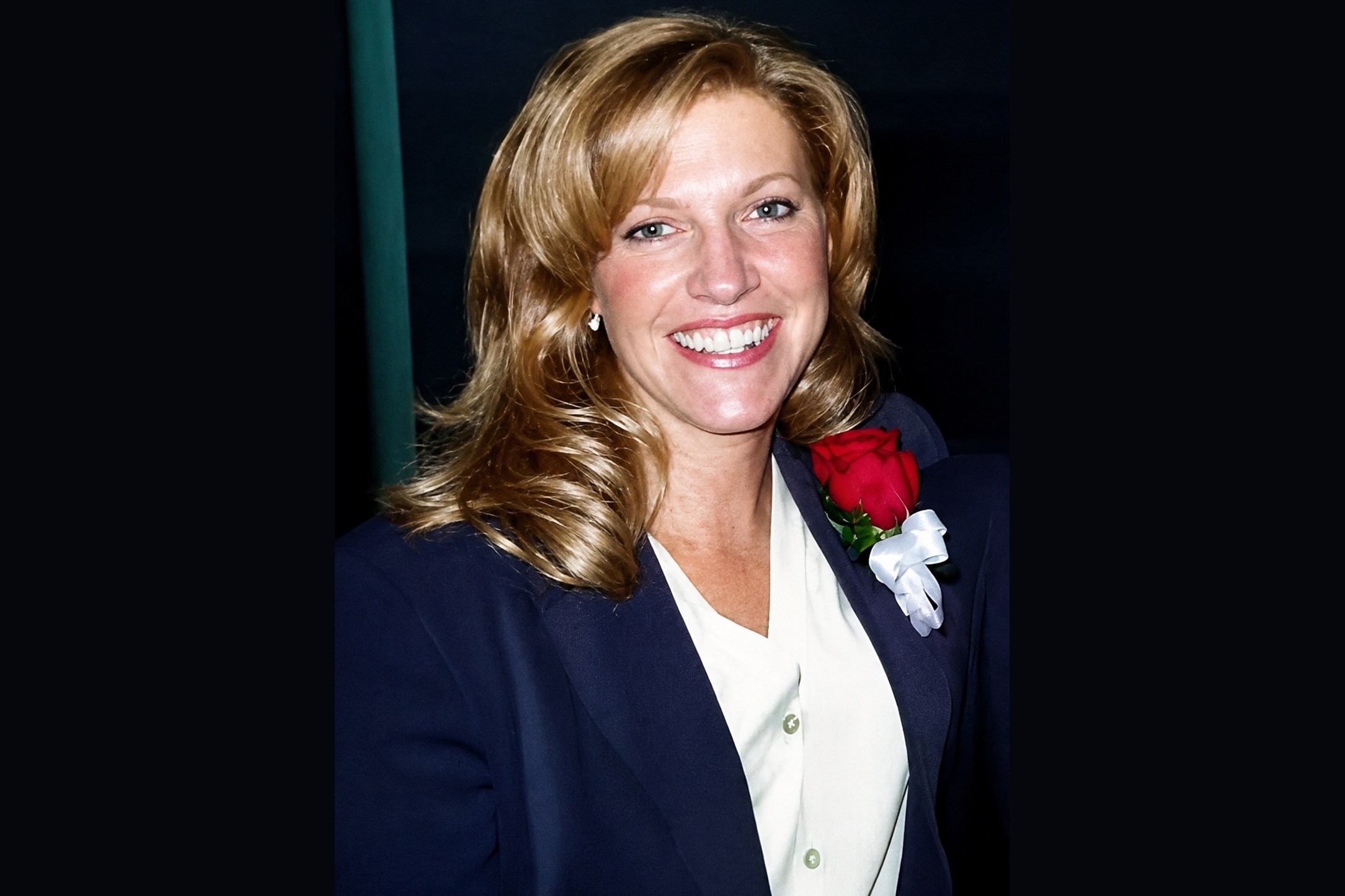 Woman smiling, wearing a dark blazer, white blouse, and a red rose boutonnière on her lapel.
