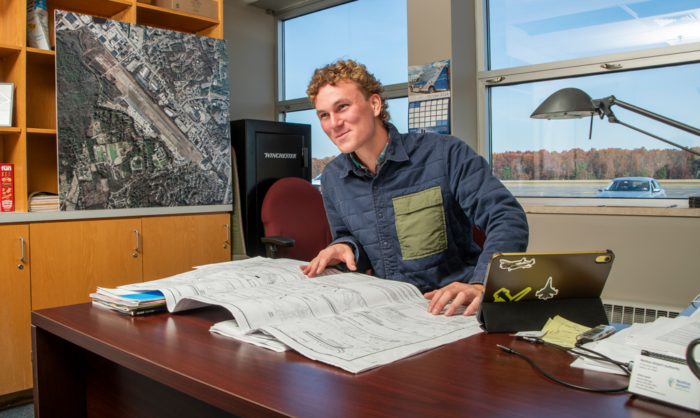 Man in an office reviews architectural plans at his desk, with maps and papers in the background.