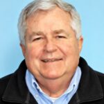 Smiling older man with gray hair in a blue shirt and black jacket, exuding leadership in front of a light blue background.