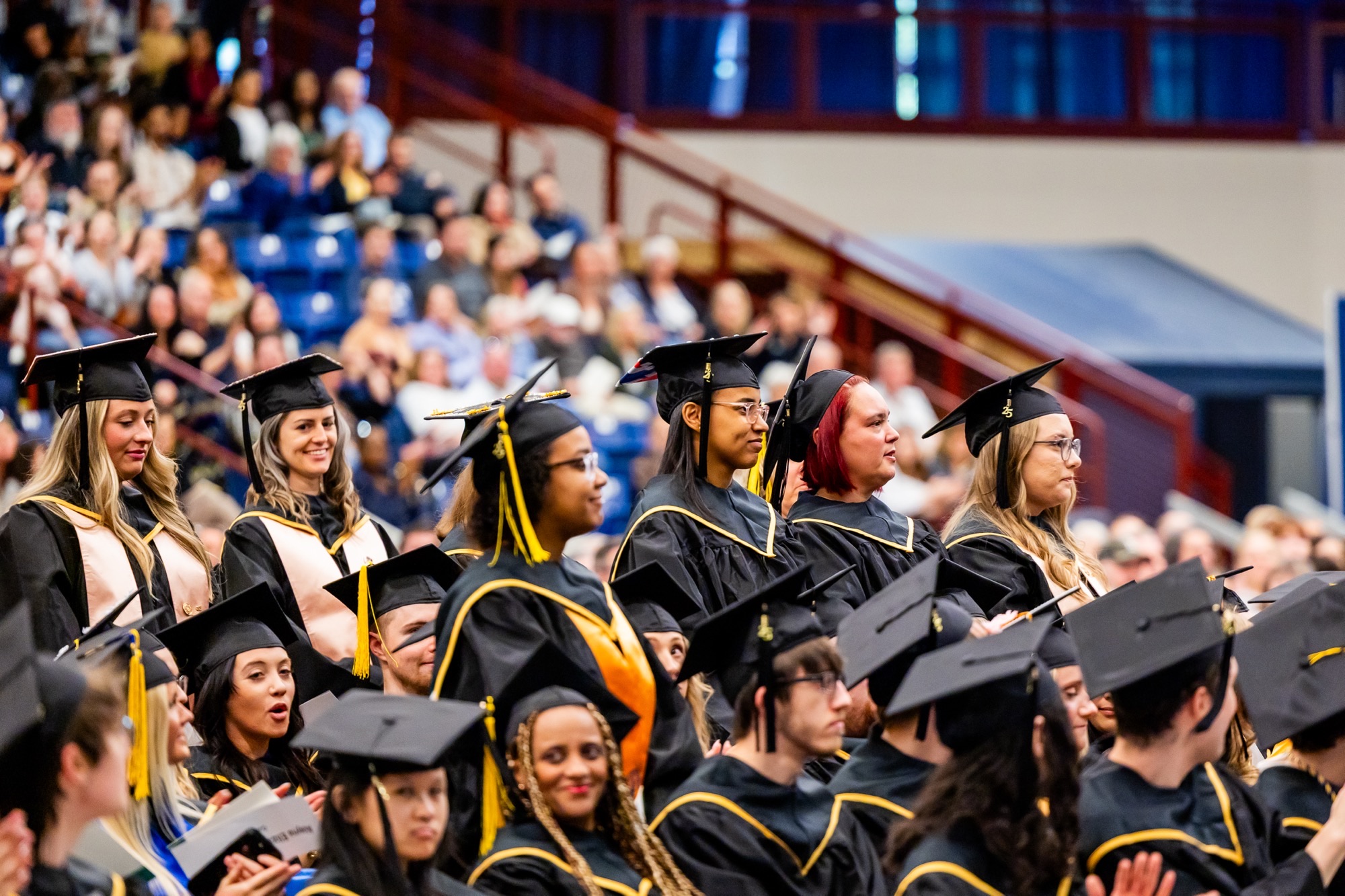 Graduates in caps and gowns sitting in an arena during a commencement ceremony.