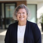 A smiling middle-aged woman in a dark cardigan and white top, who invites you to give to River Valley Community College.